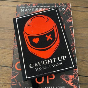 May include: A hardcover book and a black book cover with the title "CAUGHT UP" by Navessa Allen. The cover features a red helmet design with a heart and an X. The book is a "DARKNESS NOVEL".