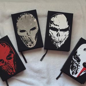 May include: Four black notebooks with skull designs. One notebook has a silver mask, another a white skull, a third a red skull, and the last a white skull with a red stripe. Each notebook has a black ribbon bookmark.