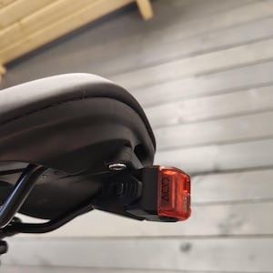 May include: A black bicycle saddle with a red and black ACID brand tail light attached. The tail light is rectangular and has the ACID logo printed on the side. The saddle is mounted on a black metal frame.