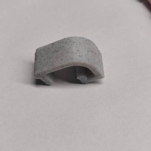 May include: A small, grey, plastic clip with a speckled texture. The clip is shaped like a C and has a rounded edge.