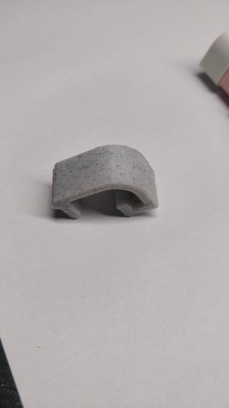 May include: A small, grey, plastic clip with a speckled texture. The clip is shaped like a C and has a rounded edge.