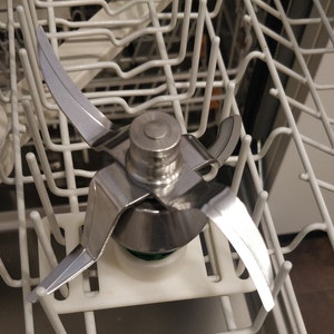 May include: A silver blender blade with three arms and a central base. The blade is in a dishwasher rack.