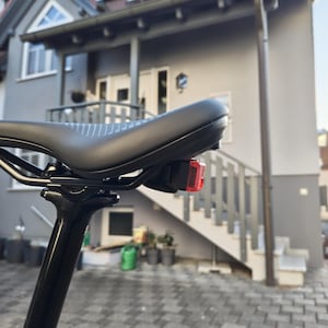 May include: A black bicycle saddle mounted on a black seat post, featuring a smooth matte finish with subtle white line patterns on the rear. A small, rectangular red safety light is securely attached to the underside of the saddle frame.