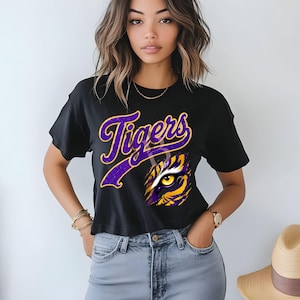 May include: A black short-sleeved t-shirt featuring the word Tigers in purple script with a yellow outline. Below the text is a graphic of a tiger eye surrounded by purple and yellow fur patterns. The shirt has a crew neckline and a straight hem.