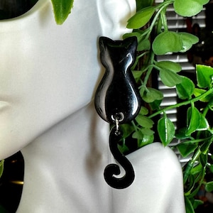 May include: Black cat dangle earrings with a curled tail. The earrings are made of a smooth, black material and are hanging from a mannequin's ear.