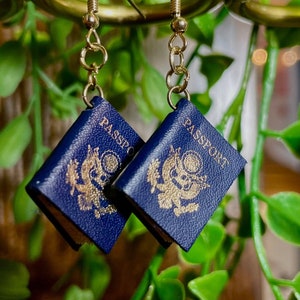 May include: A pair of gold earring hooks with blue passport charm pendants. The charms are miniature replicas of a US passport with the words "PASSPORT" and "PASSE" visible.