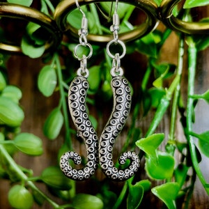 May include: Silver dangle earrings featuring a detailed octopus tentacle design. The earrings are hanging from a silver hook.