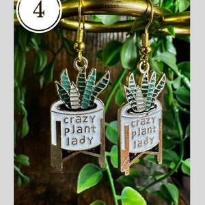May include: A pair of gold earrings with a white pot and green plant design. The pot has the text "crazy plant lady" on it.