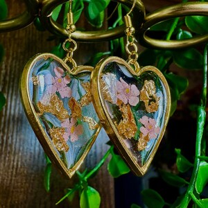 May include: A pair of gold heart-shaped earrings with a clear resin filled with dried pink flowers and gold leaf.