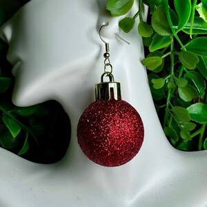 May include: Red glitter Christmas ornament earrings with gold accents. The earrings are hanging from a mannequin's ear.