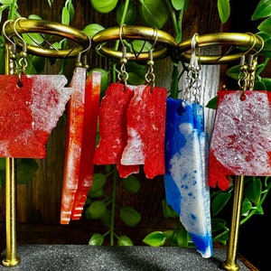 May include: Earrings in the shape of U.S. states, displayed on a gold stand. The earrings are red, white, and blue, with a marbled effect. The earrings are hanging from gold-colored hooks.
