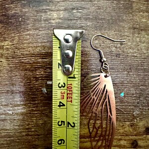 May include: A pair of copper-colored earrings shaped like butterfly wings. The earrings are measured against a yellow tape measure, showing they are approximately 2 inches long.