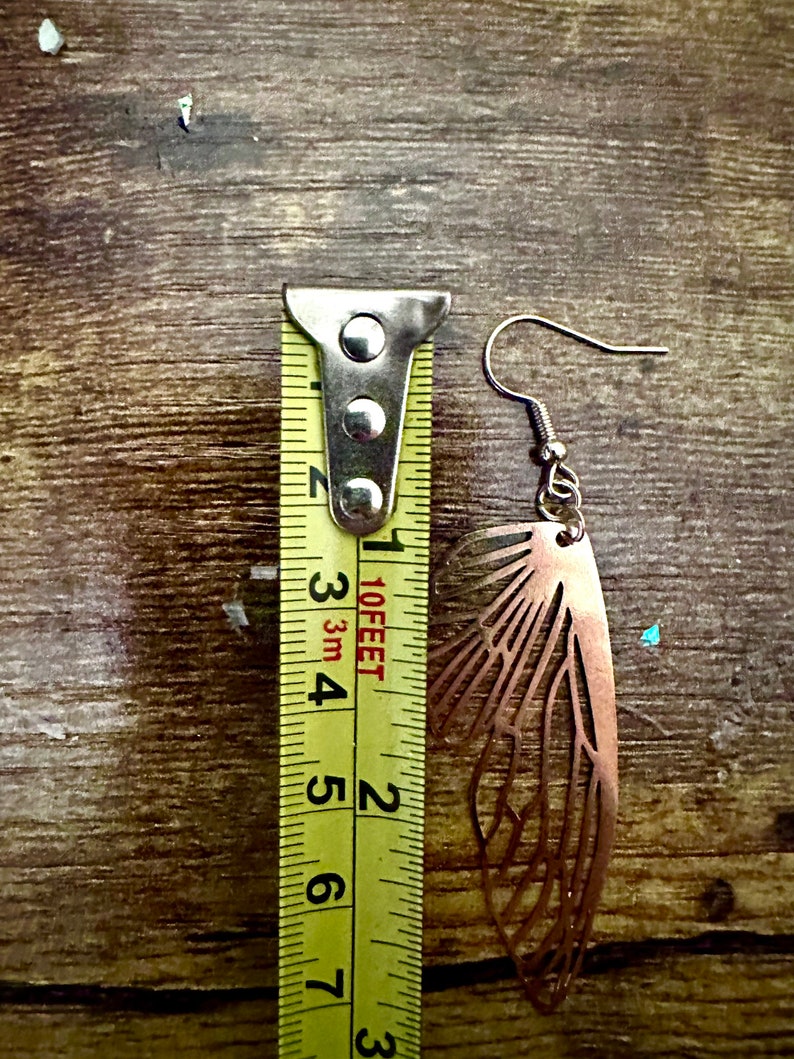 May include: A pair of copper-colored earrings shaped like butterfly wings. The earrings are measured against a yellow tape measure, showing they are approximately 2 inches long.