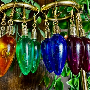 May include: A set of six colourful lightbulb earrings. The earrings are made of glass and have a gold-coloured metal hook. The lightbulbs are in various colours, including orange, green, blue, and purple.