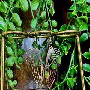 May include: Gold-toned earrings shaped like butterfly wings with intricate cutouts. The earrings are displayed on a gold stand.