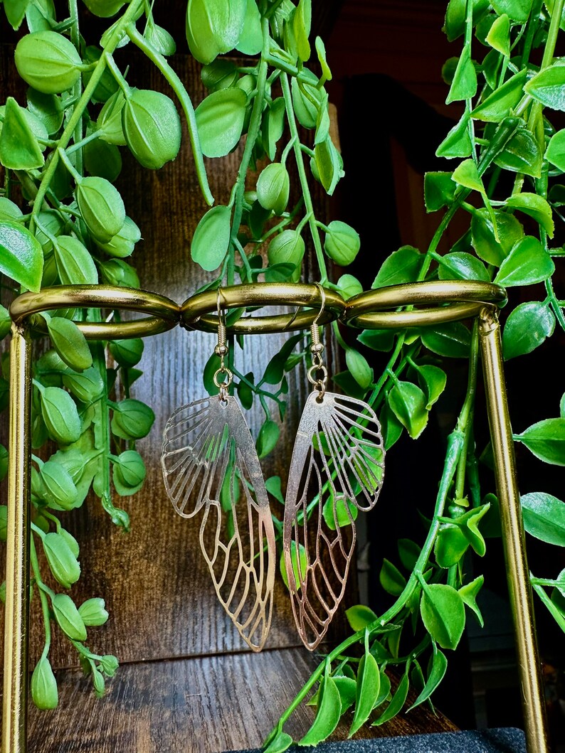 May include: Gold-toned earrings shaped like butterfly wings with intricate cutouts. The earrings are displayed on a gold stand.
