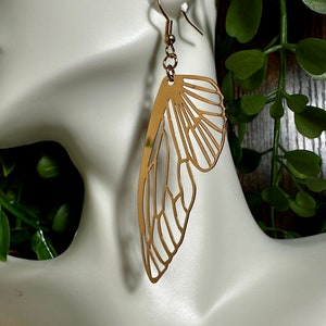 May include: Gold-toned earrings shaped like a butterfly wing. The earrings are delicate and intricate, with a detailed design.