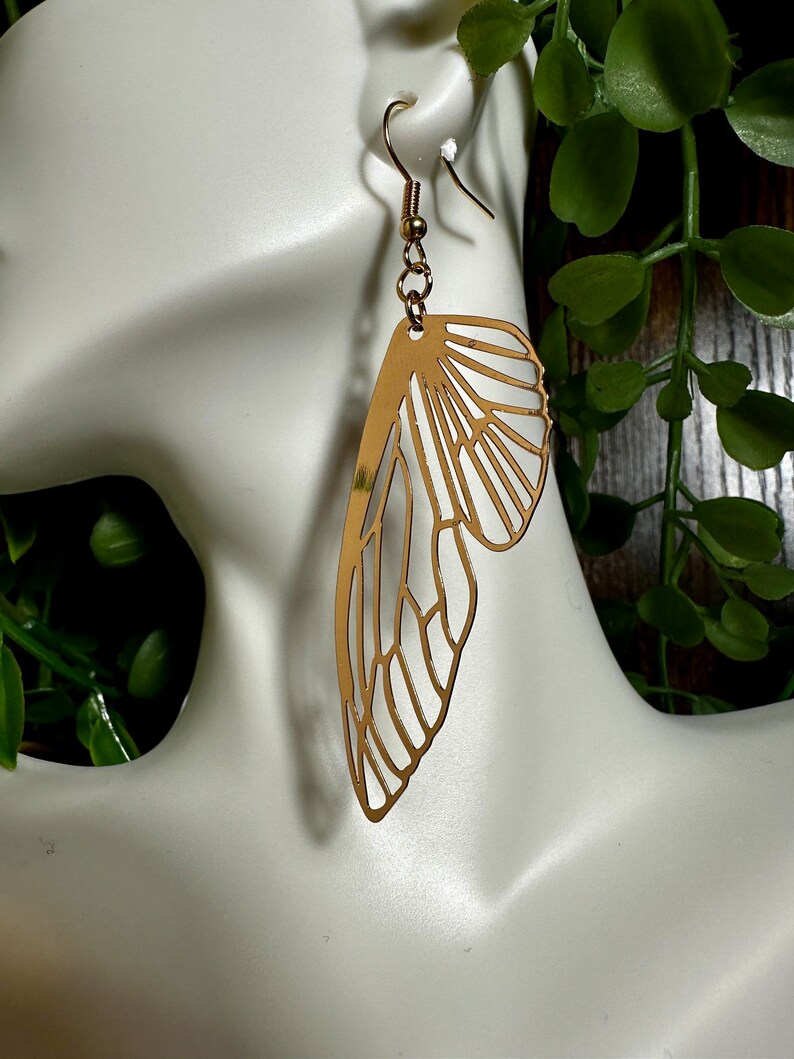 May include: Gold-toned earrings shaped like a butterfly wing. The earrings are delicate and intricate, with a detailed design.