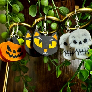 Può includere: Un set di orecchini a tema Halloween con un gatto nero, una zucca e due teschi bianchi. Gli orecchini sono esposti su un supporto dorato.