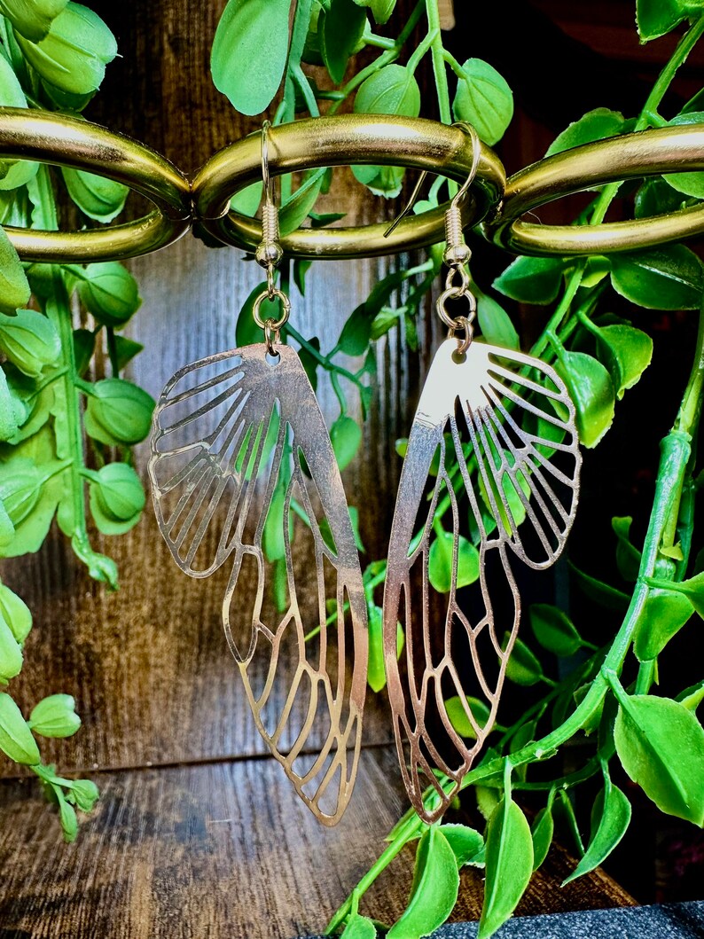 May include: A pair of gold-toned earrings shaped like butterfly wings. The earrings are hanging from a gold-toned chain.