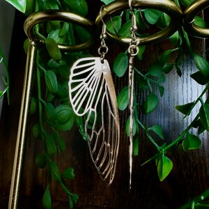 May include: Gold tone earrings with a delicate butterfly wing design and a long, thin, straight dangle. The earrings are hanging on a gold metal earring stand.