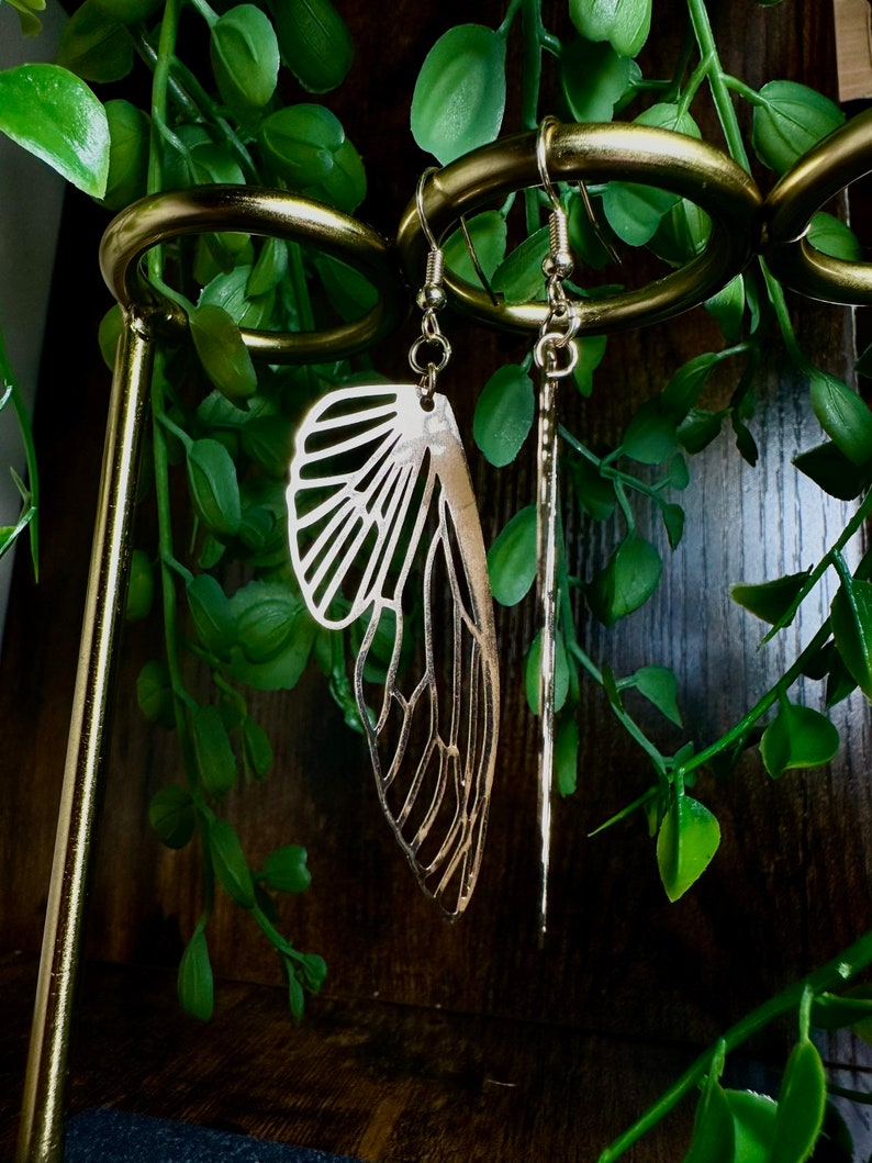 May include: Gold tone earrings with a delicate butterfly wing design and a long, thin, straight dangle. The earrings are hanging on a gold metal earring stand.