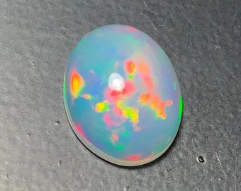 Puede incluir: Una piedra preciosa de ópalo cabujón de forma ovalada con una base blanca lechosa translúcida y destellos vibrantes de rojo, naranja, amarillo y verde que juegan en toda la piedra. La superficie es lisa y pulida con un perfil de cúpula alta.