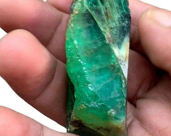 Può includere: Un cristallo di smeraldo grezzo e allungato caratterizzato da una tonalità verde vibrante con sezioni traslucide, fratture interne visibili e inclusioni minerali scure su tutta la superficie naturale sfaccettata della pietra.