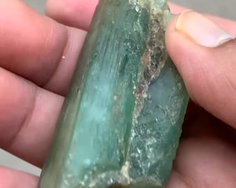 May include: A raw, elongated crystal specimen with a translucent, pale green hue. The mineral features a columnar structure with vertical striations along its length, interspersed with rough, textured patches and earthy brown mineral inclusions.