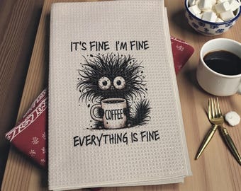 May include: A cream-colored kitchen towel with a textured waffle weave. The towel features a cartoon monster holding a coffee mug with the word "COFFEE" and the text "IT'S FINE I'M FINE EVERYTHING IS FINE".