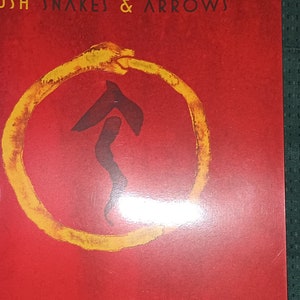 May include: Album cover with a red background and the text "RUSH SNAKES & ARROWS" at the top. A yellow circle surrounds a black and red arrow-like symbol. The album art has a grunge, textured appearance.