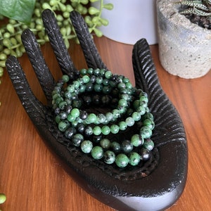 May include: A set of five green and black gemstone bracelets stacked on a black hand-shaped display. The bracelets are made of small, round beads.