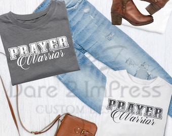 May include: Two folded t-shirts with the text "Prayer Warrior" in a white and gray design on a white background and a gray background. The shirts are displayed with a brown leather purse, blue jeans, and brown boots.