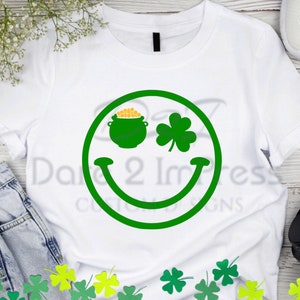 May include: White t-shirt with a green smiley face design. The smiley face has a pot of gold with a shamrock on top of it.