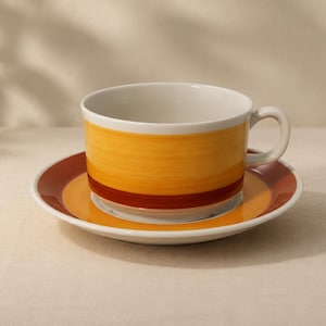 May include: A ceramic teacup and saucer set. The cup has a white interior and a yellow and orange exterior with a dark red band. The saucer has a matching color scheme. The set is designed for serving hot beverages.