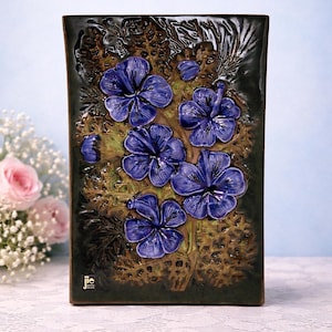 May include: A rectangular ceramic vase featuring a dark green base with a raised design of blue flowers. The flowers have multiple petals and are set against green foliage. The vase has a glossy finish and a small logo.