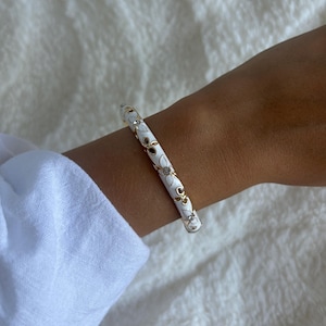 Peut inclure: Un bracelet jonc fin avec une finition en émail blanc, agrémenté de motifs floraux en métal doré et de petits cristaux transparents incrustés. Le bracelet présente un profil lisse et arrondi, conçu pour être porté au poignet comme accessoire.