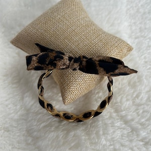 Pode incluir: Uma pulseira dourada com um laço com estampa de leopardo. A pulseira apresenta um design tipo corrente com elos dourados e tecido com estampa de leopardo. O laço é amarrado na parte superior da pulseira.