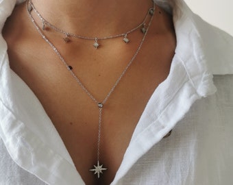 Puede incluir: Un collar plateado de varias capas con detalles de colgantes de estrella y cristal. La capa superior presenta pequeños dijes de estrella, mientras que la capa inferior tiene un colgante de estrella fugaz. El collar se usa sobre una camisa blanca abotonada.