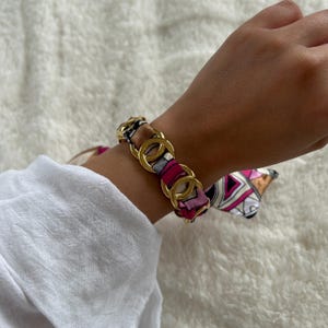 Peut inclure: Un bracelet à chaîne doré avec un motif d'écharpe en soie colorée. Le bracelet présente de grands maillons circulaires imbriqués. Le motif de l'écharpe comprend des nuances de rose, de marron et de blanc, ajoutant une touche d'élégance.