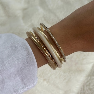 Peut inclure: Plusieurs bracelets dorés et crème empilés sur un poignet. Les bracelets comprennent une bande dorée texturée, une bande crème et une bande dorée avec de petits embellissements scintillants. Une manche de chemise blanche est visible en arrière-plan.