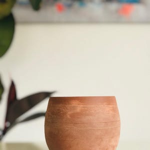 May include: A small, round, terracotta-colored planter with horizontal etched lines. The unglazed clay pot has a slightly curved shape and is suitable for small plants. The background is blurred, with hints of green foliage and abstract art.