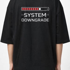 Puede incluir: Camiseta negra con el texto "::SYSTEM:: DOWNGRADE" en blanco. Encima del texto hay un gráfico de barra de carga roja y blanca con una flecha. La camiseta tiene mangas cortas y un corte holgado.