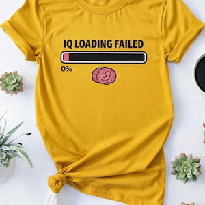 May include: Mustard yellow t-shirt with rolled sleeves. The shirt features the text "IQ LOADING FAILED" above a loading bar at 0% and a pink brain graphic. The shirt is knotted at the bottom.