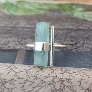 Puede incluir: Un anillo de plata con una piedra de aguamarina azul claro y una barra de plata en la parte superior. El anillo está sobre un fondo marrón. SilverArtisanGift