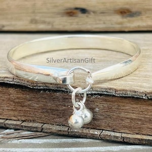 May include: A silver bangle bracelet with a small loop and two dangling silver ball charms. The bracelet is smooth and shiny, reflecting the light. The bracelet is displayed on a wooden surface.
