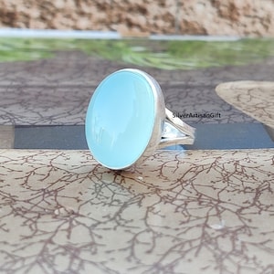 May include: A silver ring with a large, oval, pale blue gemstone. The gemstone is set in a polished silver band. The ring is displayed on a patterned surface. The text "SilverArtisanGift" is visible.
