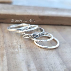 Könnte beinhalten: Eine Auswahl an Silberringen mit verschiedenen Texturen und Designs. Ein Ring hat ein Blumenmuster, andere haben gehämmertes oder glattes Finish. Die Ringe werden auf einer Holzoberfläche präsentiert, mit dem Text "SilverArtisanGift" sichtbar.
