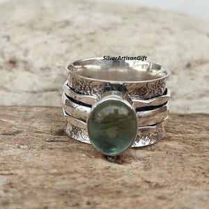 May include: A silver ring featuring a green gemstone set in a textured band. The ring is crafted from multiple bands stacked on top of each other.