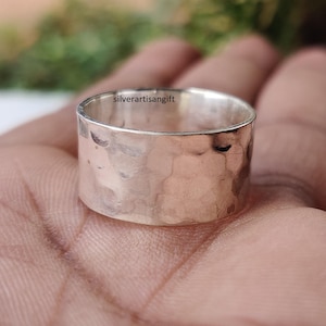 Puede incluir: Un anillo ancho de plata martillada. El anillo tiene una superficie texturizada con una serie de pequeñas hendiduras. El texto "silverartisangift" es visible. El anillo se sostiene en una mano.